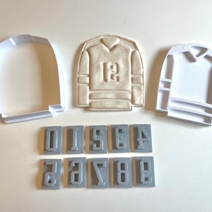 May include: A set of cookie cutters for making hockey jerseys. The set includes a large jersey cutter, a smaller jersey cutter, and ten number cutters. The number cutters are 0, 1, 2, 3, 4, 5, 6, 7, 8, and 9.