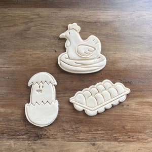 May include: Three cookie cutters made of white dough, shaped like a hen in a nest, a chick in an eggshell, and a carton of eggs.