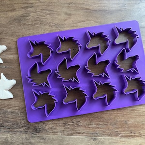 May include: Purple plastic cookie cutter set with 12 unicorn shapes. The cutters are arranged in a grid pattern. Two white unicorn cookies are shown next to the cutters.