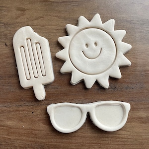 May include: Three white cookie cutters shaped like a popsicle, sunglasses, and a smiling sun.