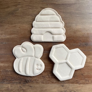 May include: Three white cookie cutters in the shapes of a beehive, a bee, and a honeycomb. The beehive has horizontal lines and a small opening. The bee has a smiling face and striped body. The honeycomb has three connected cells.