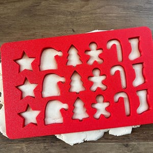 May include: Red plastic Christmas cookie cutter with various shapes including stars, Santa hats, Christmas trees, candy canes, and boots. The cutter is on top of a white dough.