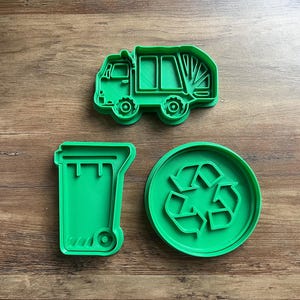May include: Green plastic cookie cutters shaped like a garbage truck, a recycling symbol, and a trash can.