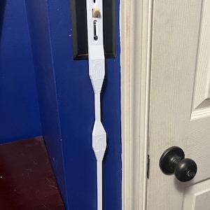 May include: White plastic extension for a light switch, attached to a wall with a blue painted surface. The extension is shaped like a long, thin rectangle with a rounded end. It has a series of raised, decorative patterns along its length.