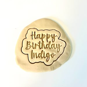 May include: A close-up of a cookie cutter with the words "Happy Birthday Indigo" in a cursive font.
