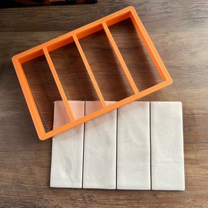 May include: An orange plastic mold with four rectangular slots, used for making soap bars. The mold is placed on top of four white soap bars.