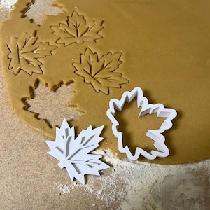 May include: White plastic maple leaf cookie cutters on a brown surface with a rolled out sheet of cookie dough. The cookie dough has been cut out with the cookie cutters to create maple leaf shapes.