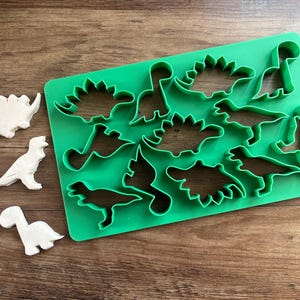 May include: Green plastic dinosaur cookie cutters on a wooden surface. The cutters include a variety of dinosaur shapes, including a stegosaurus, triceratops, and brachiosaurus.