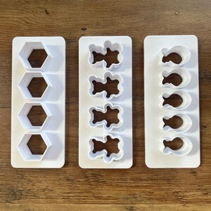May include: Three white plastic cookie cutters with different shapes. The first cutter has four hexagonal shapes. The second cutter has four bear shapes. The third cutter has four fish shapes.