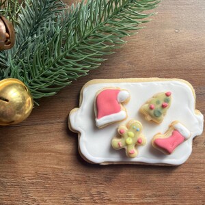 May include: A white plate with four Christmas-themed sugar cookies. The cookies are decorated with red and green icing and feature a Santa hat, a Christmas tree, a snowman, and a stocking.