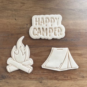 May include: Three cookie cutters for camping themed cookies. The cutters are white and feature a campfire, a tent, and the words "Happy Camper".