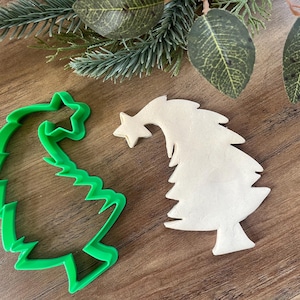 May include: A green plastic Christmas tree cookie cutter and a white cookie in the shape of a Christmas tree.