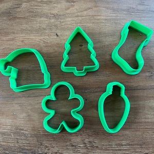 May include: Five green plastic Christmas cookie cutters: a stocking, a Christmas tree, a gingerbread person, a Santa hat, and a light bulb.