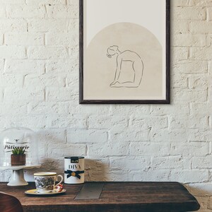 May include: A framed print of a line drawing of a person in a yoga pose. The drawing is on a beige background with a brown circle behind the person. The frame is black.