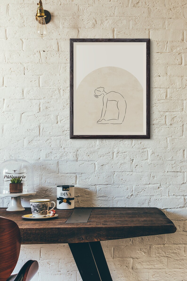 May include: A framed print of a line drawing of a person in a yoga pose. The drawing is on a beige background with a brown circle behind the person. The frame is black.