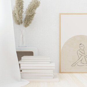 May include: A framed print with a minimalist line drawing of a person sitting in a circle on a beige background. The frame is light wood and the print is hanging on a white wall.