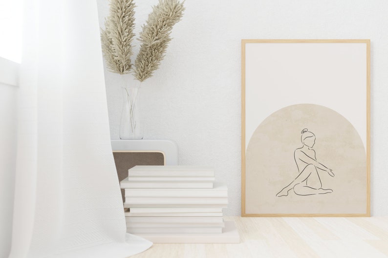 May include: A framed print with a minimalist line drawing of a person sitting in a circle on a beige background. The frame is light wood and the print is hanging on a white wall.