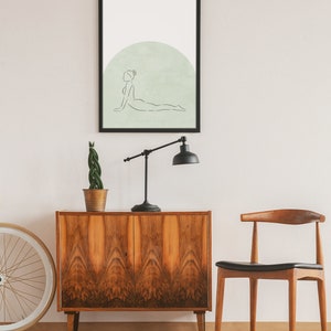 May include: A black framed print of a line drawing of a person in a yoga pose on a light green background. The print is hanging on a white wall above a wooden cabinet and a wooden chair.