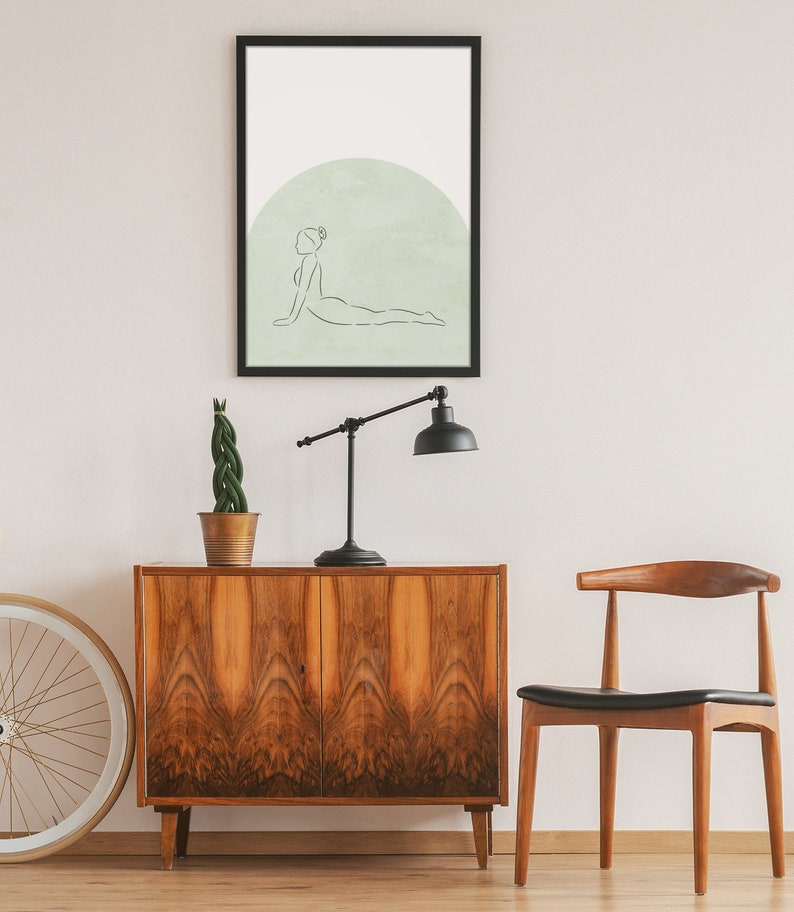 May include: A black framed print of a line drawing of a person in a yoga pose on a light green background. The print is hanging on a white wall above a wooden cabinet and a wooden chair.