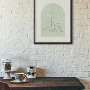 May include: A framed print of a line drawing of a person doing a yoga pose in front of a green arch. The print is hanging on a white brick wall.