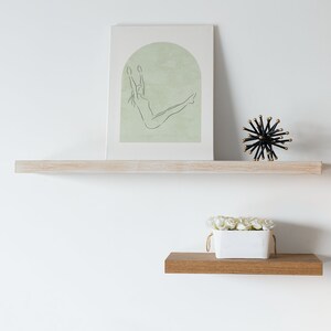 May include: A white shelf with a framed print of a line drawing of a woman in a yoga pose. The print has a green background. A black and gold decorative object and a white planter with white flowers are on the shelf.