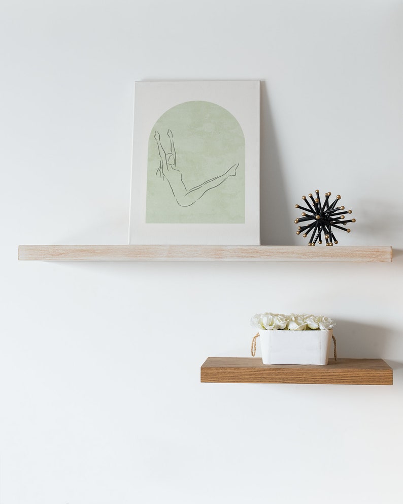 May include: A white shelf with a framed print of a line drawing of a woman in a yoga pose. The print has a green background. A black and gold decorative object and a white planter with white flowers are on the shelf.
