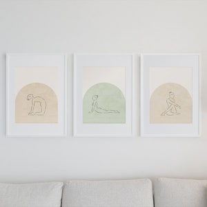 May include: Three white framed prints with minimalist line drawings of people in yoga poses on a beige background. Each print features a different pose and a subtle color gradient in the background.