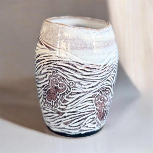 May include: A handmade ceramic tumbler with a unique design. The upper portion is white, transitioning to a brown and white striped pattern with brown animal figures. The base is black, and the interior is white.