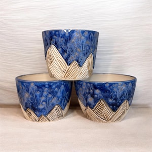 May include: Three ceramic bowls with a mountain design. The bowls have a blue glaze with a textured appearance, and the mountain range is carved in a cream color. The bowls are stacked on top of each other.