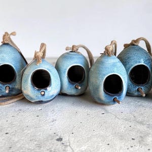 May include: Seven teardrop-shaped ceramic birdhouses in a mottled blue glaze. Each house features a circular entrance hole, a small wooden perch, and a thick jute rope loop attached to the top for hanging. The surfaces show variations in blue tones.