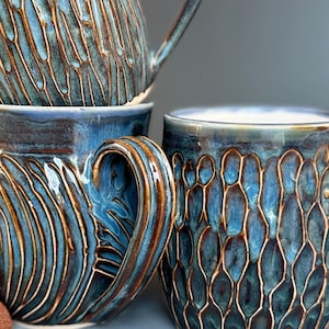 May include: Close-up of several handmade ceramic mugs. The mugs are glazed in shades of blue and brown, with textured patterns. The handles are curved and the mugs are stacked, showcasing the craftsmanship and design.
