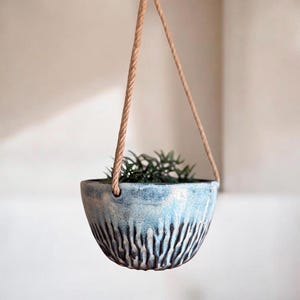 May include: A hanging ceramic planter with a light blue glaze and vertical textured lines. The planter is suspended by a natural rope and contains green foliage. A decorative piece for indoor or outdoor use.