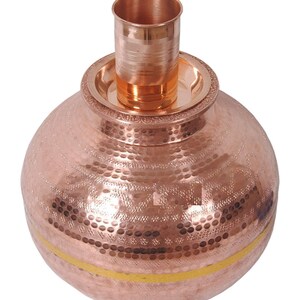 May include: A copper water pot with a hammered finish and a gold band around the middle. The pot has a small copper cup on top.