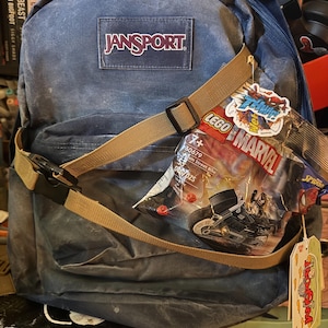 May include: A blue Jansport rucksack with a tan strap and buckle. A Lego Marvel set is attached to the rucksack. The rucksack has the word "JANSPORT" on the front.