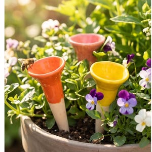 May include: Close-up of a terracotta flower pot filled with green plants and colorful pansies. Three ceramic plant watering spikes in orange, pink, and yellow are inserted into the soil. A bee is perched on the orange spike.