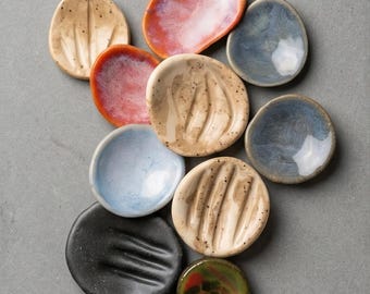 May include: A collection of ten small, round, and oval ceramic dishes in various colors including black, blue, beige with speckles, orange, and green. Some dishes feature grooved textures, while others have smooth, glazed surfaces. Text reads: A complete tactile experience.