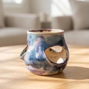 May include: A handcrafted ceramic vessel with a unique design. The piece features a cylindrical shape with two large circular openings and a small spout. The exterior is painted with a blend of blue, purple, and pink watercolor-like hues, creating a marbled effect. The vessel sits on a wooden surface.