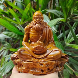 May include: A wooden sculpture of a seated Buddha with a long beard and a serene expression. The sculpture is carved in a detailed style and has a warm, golden brown colour.
