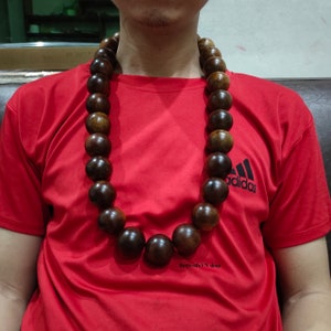 Könnte beinhalten: Eine lange braune Holzperlenkette mit großen runden Perlen. Die Kette wird von einer Person getragen, die ein rotes Shirt mit dem Wort "adidas" auf der Vorderseite trägt.