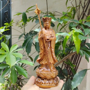 Könnte beinhalten: Eine Holzstatue einer Gottheit mit einer Krone und einem Stab, der mit einer Blume gekrönt ist. Die Statue steht auf einem Lotussockel. Die Statue ist braun und hat eine natürliche Holzoptik.