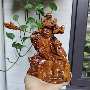 Op de afbeelding: Een gedetailleerd houten beeldhouwwerk van een krijgerfiguur die een speer vasthoudt. De figuur staat op een voetstuk met een drakenontwerp. Het beeldhouwwerk is van een rijke bruine kleur.