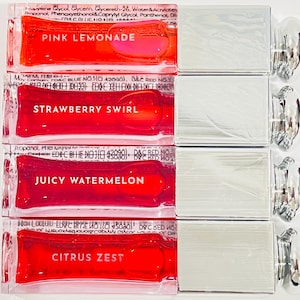 Könnte beinhalten: Vier rechteckige Lipgloss-Behälter mit den Geschmacksrichtungen Pink Lemonade, Strawberry Swirl, Juicy Watermelon und Citrus Zest. Jeder Behälter hat ein klares, glänzendes Aussehen mit dem Geschmacksnamen in weißem Text. Der Lipgloss hat eine leuchtend rote Farbe.
