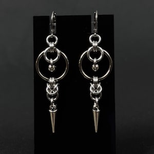 Puede incluir: Un par de pendientes plateados con un diseño geométrico. Cada pendiente presenta múltiples anillos conectados, pequeñas cuentas y una punta en forma de cono en la parte inferior. Los pendientes se muestran sobre un fondo negro.