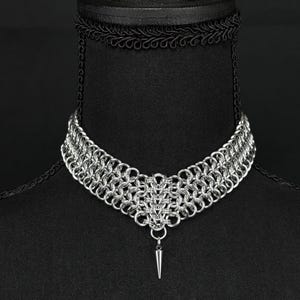 May include: A silver chainmail choker necklace with a pointed pendant. The necklace is displayed on a black mannequin.