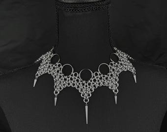 May include: A silver-toned chainmail choker necklace featuring a complex woven metal link pattern. The design includes five circular metal rings along the top edge and five downward-pointing metal spikes suspended from the bottom edge.