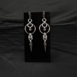 May include: A pair of silver-toned dangle earrings featuring a chainmaille design. Each earring consists of a circular hoop connected to a series of interlocking metal rings, ending in a pointed, cone-shaped metal spike charm at the bottom.