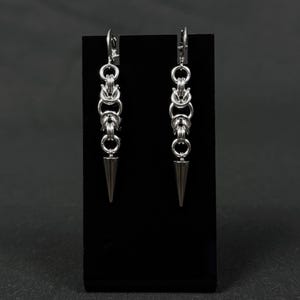 May include: A pair of silver earrings with a pointed tip and a chain-like design. The earrings are displayed on a black stand.