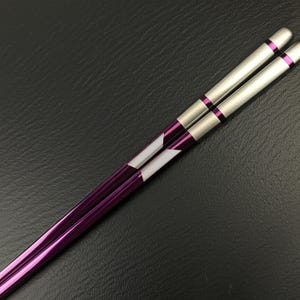 May include: A pair of purple and silver chopsticks. The chopsticks have a metallic finish with a purple base and silver tips. Each chopstick features a white and purple geometric design near the top. The chopsticks are designed for Asian cuisine.