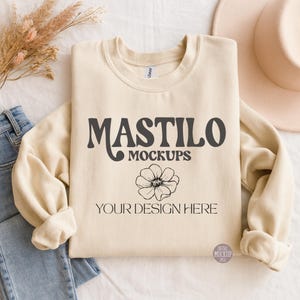 Puede incluir: Una sudadera color crema con el texto "MASTILO MOCKUPS" y "YOUR DESIGN HERE" en gris oscuro. Un gráfico floral está debajo del texto. La sudadera está doblada, con unos vaqueros azules y un sombrero en el fondo.