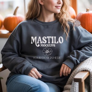 May include: A dark gray sweatshirt with the text "MASTILO MOCKUPS" in white, along with a floral graphic and the words "YOUR DESIGN HERE". The sweatshirt is worn by a person sitting in a chair, with pumpkins in the background.